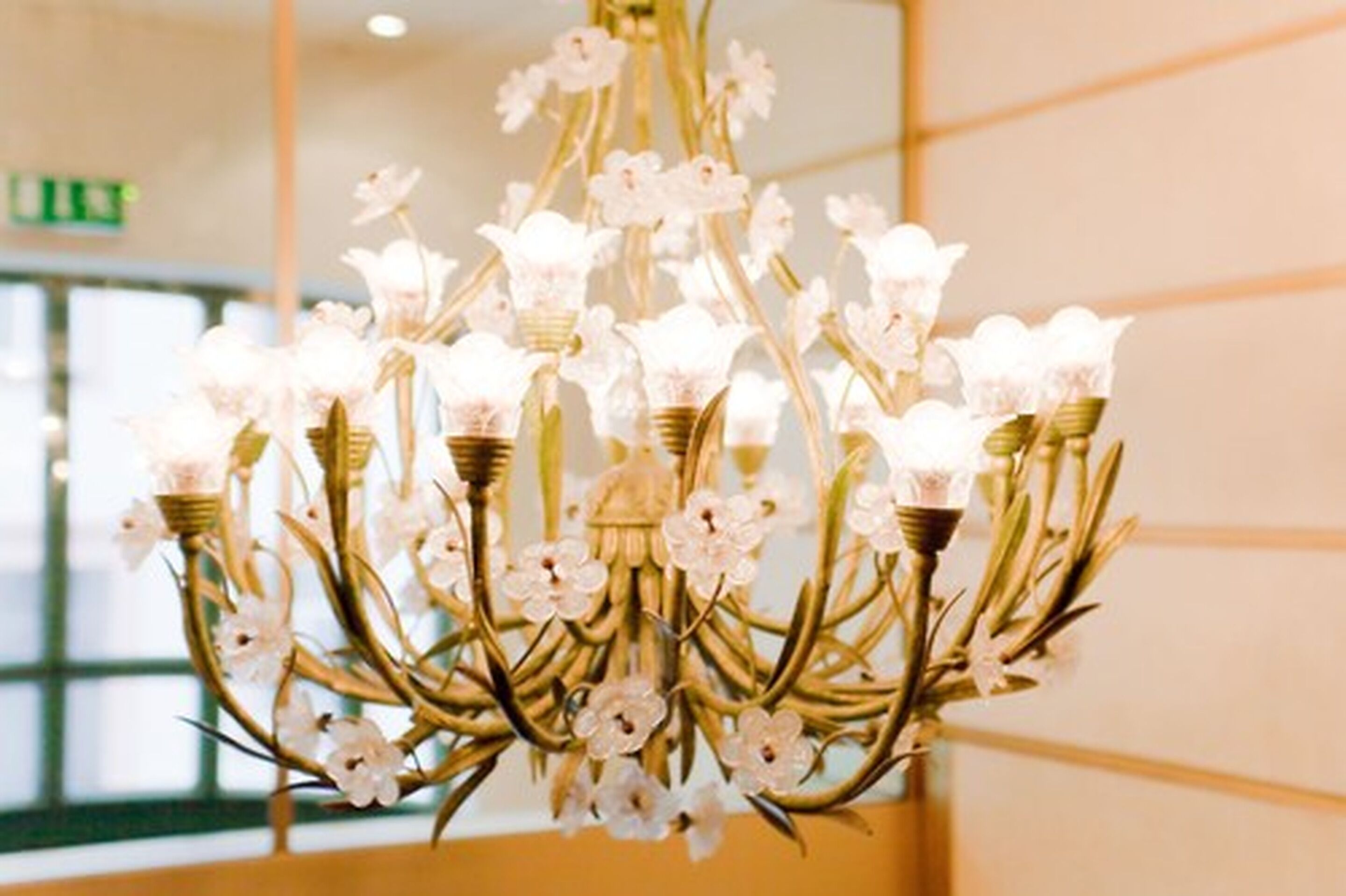 A chandelier with multiple arms and light fixtures, set in a room with a neutral color scheme and a visible exit sign.