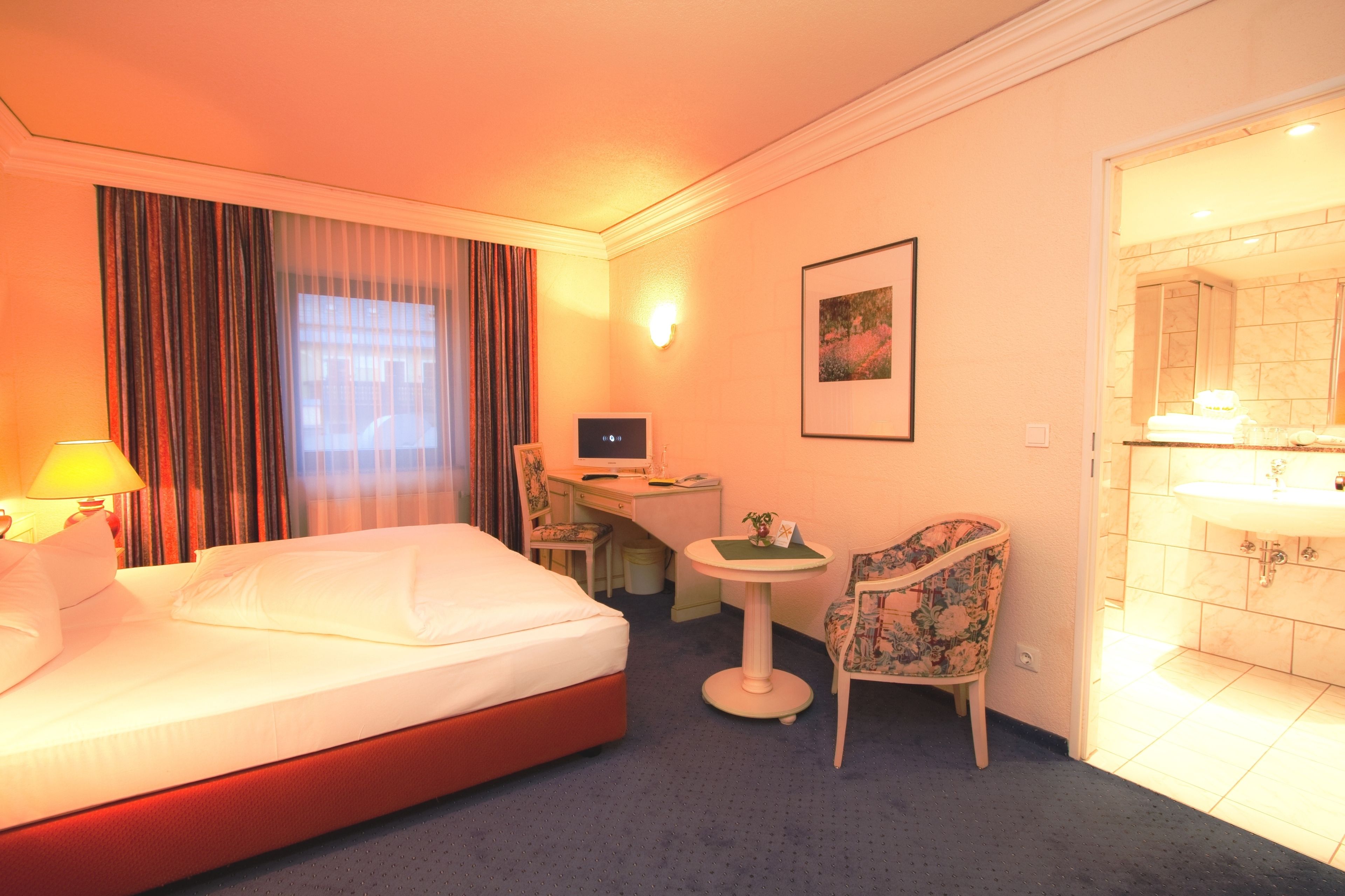 A hotel room with a bed, desk, chair, and a small table with a lamp.