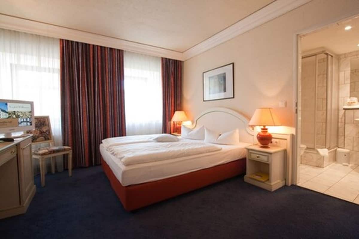 A hotel room with a bed, two bedside tables, a desk, a chair, a shower area, and curtains.