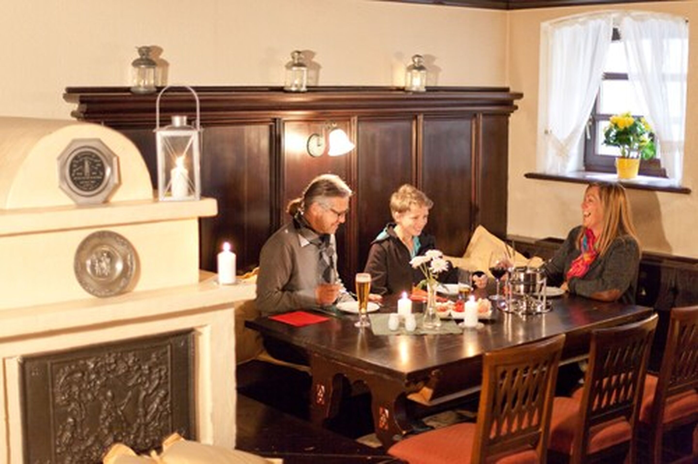 A dining room with a wooden table, chairs, and a fireplace. There are candles on the table, and a window with curtains.