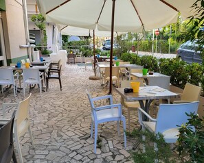 An outdoor patio area with tables and chairs, some with umbrellas, and a stone-tiled floor. - Hotel Viscount (Riccione)