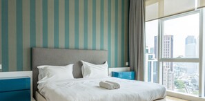 A hotel room with a large bed, striped walls, a city view, and a blue bedside table.