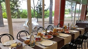 Un comedor con mesas dispuestas para el desayuno, asientos al aire libre y vistas a árboles y un edificio al fondo.