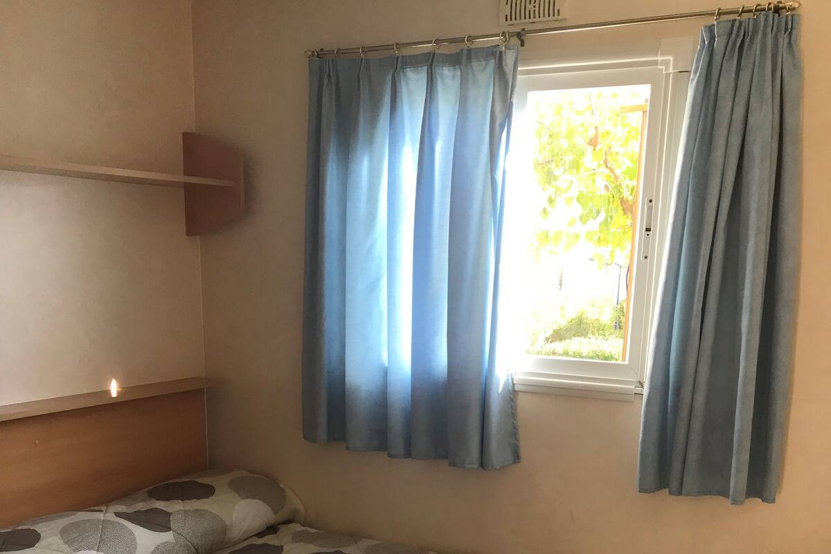 A single bed with a patterned bedspread, two rolled towels, and a window with blue curtains.