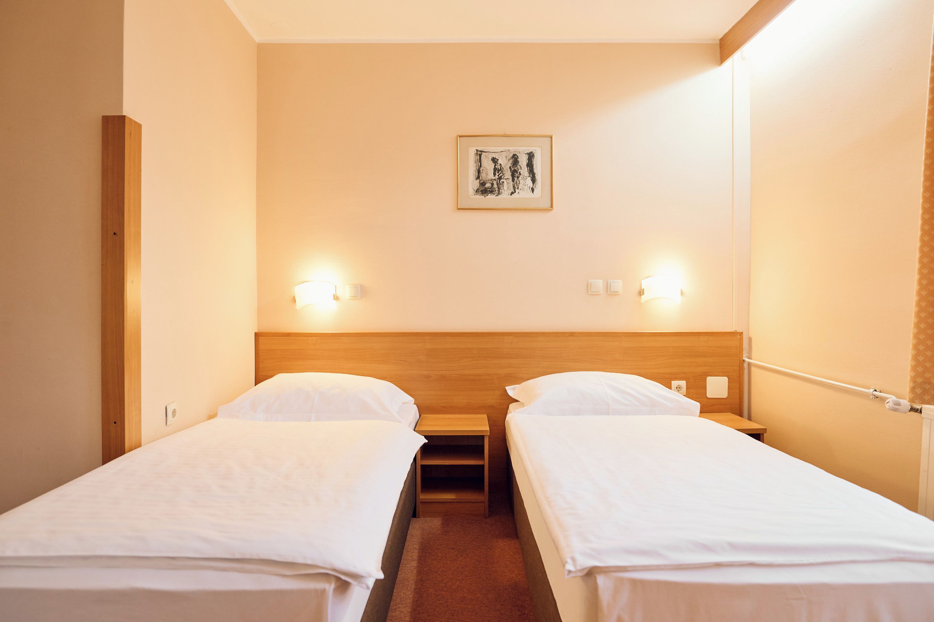 A hotel room with two single beds, a wooden headboard, a small table, and a framed artwork on the wall.