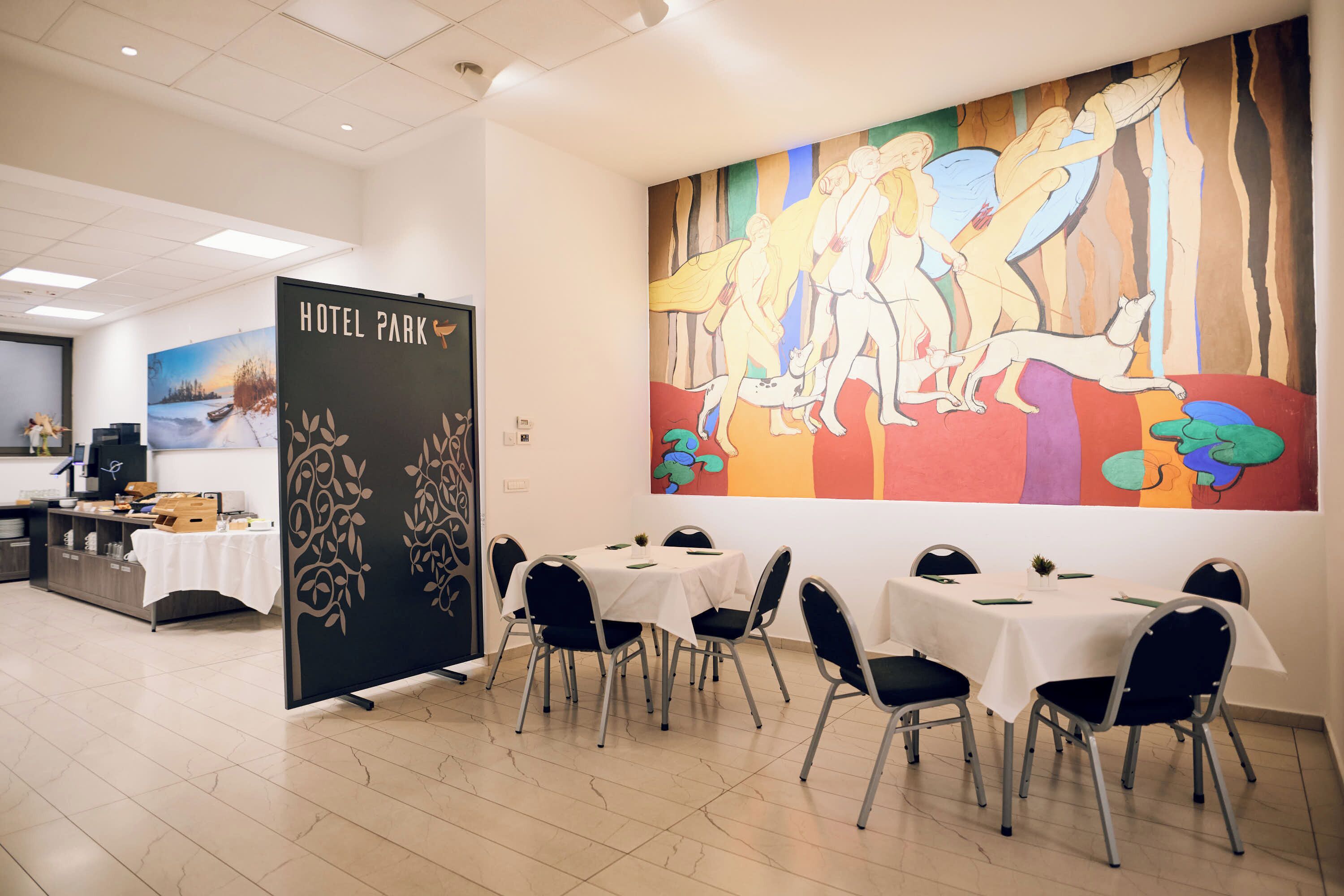 A hotel dining area with tables set for guests, a large mural on the wall, and a 'HOTEL PARK' sign.