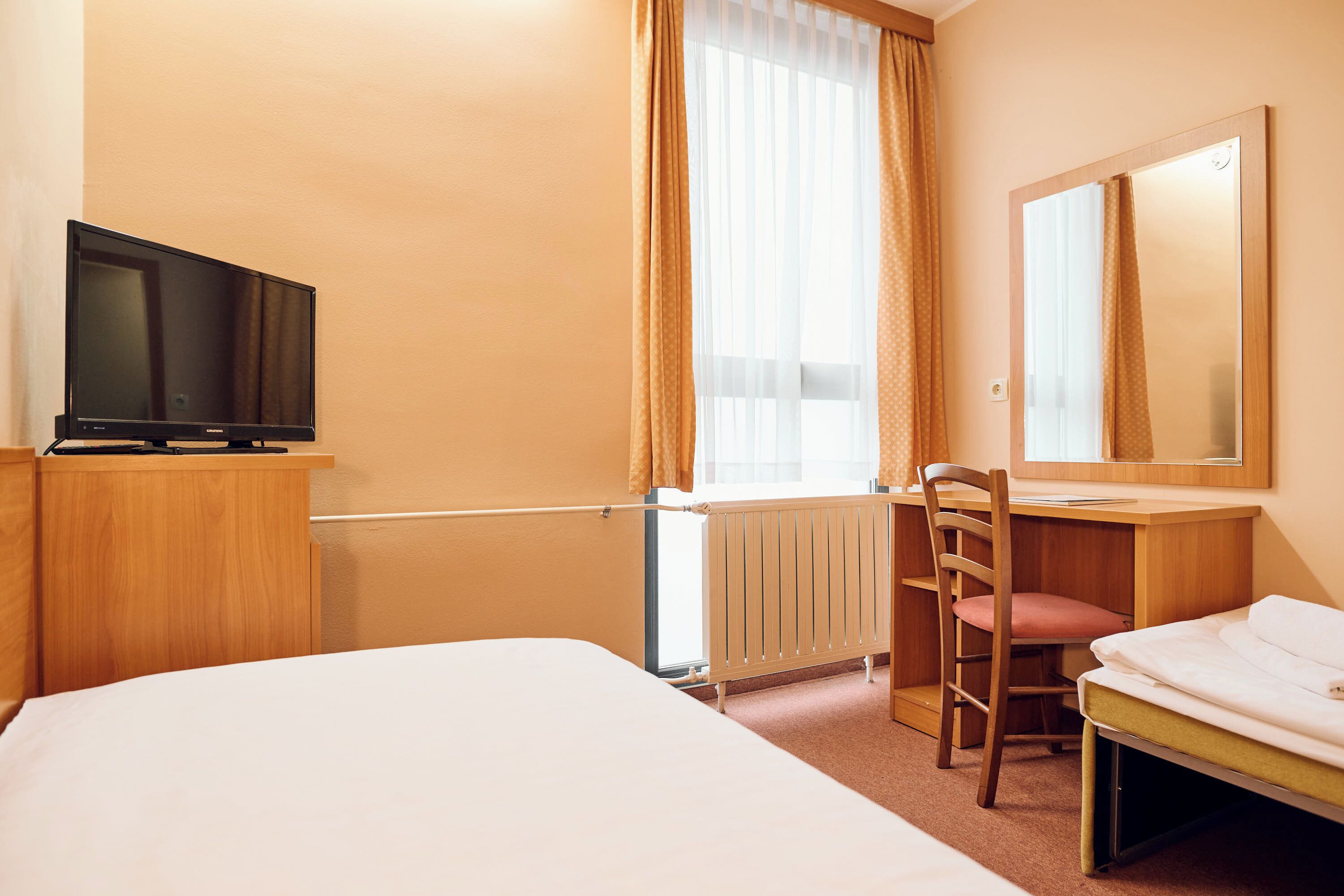 Ein Hotelzimmer mit Bett, Schreibtisch, Stuhl, Fernseher und einem Fenster mit Vorhängen.