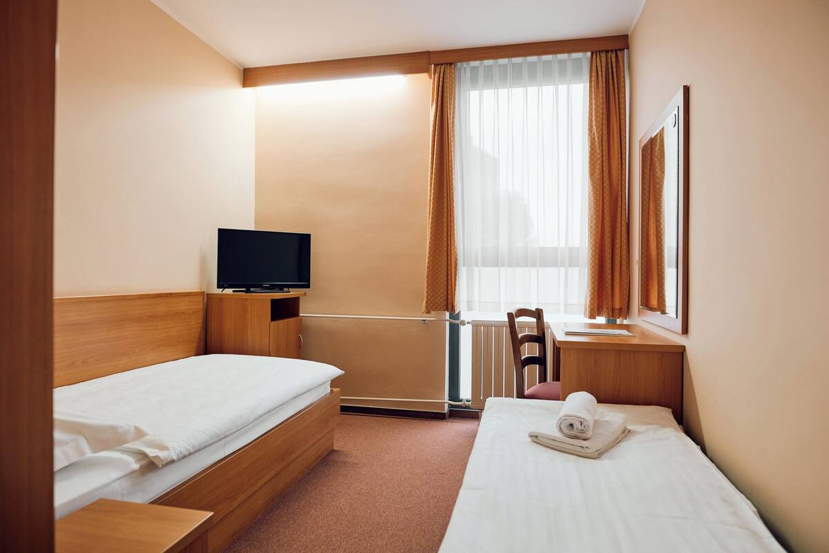A hotel room with two single beds, a wooden headboard, a small table with a television, a chair, and a window with curtains.