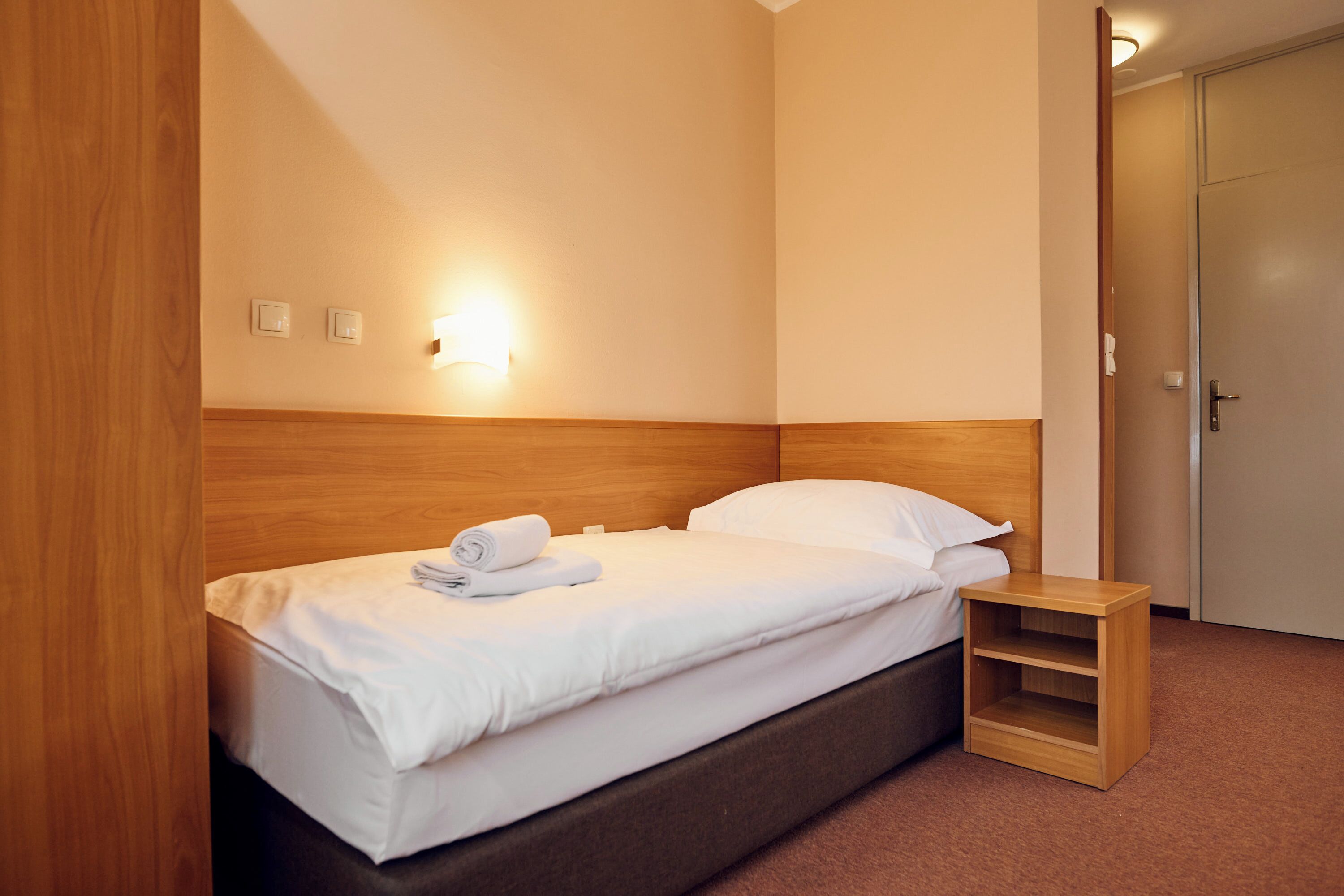 A neatly made bed with white linens and a rolled towel, a bedside table, and a wall-mounted light fixture.