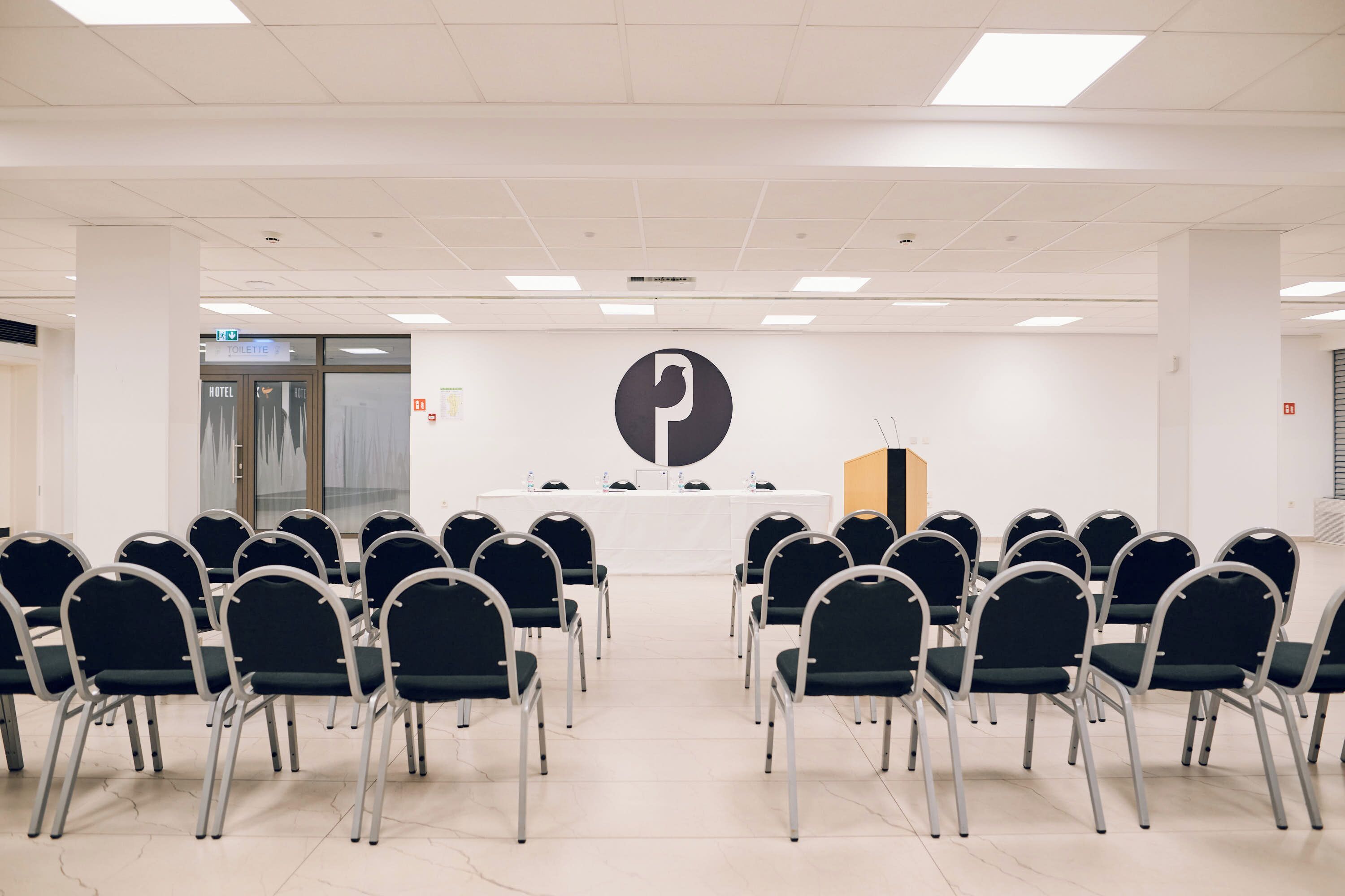 Un salón de conferencias con filas de sillas orientadas hacia un atril, un logotipo grande en la pared y una puerta con la leyenda 'TOILET' al fondo.