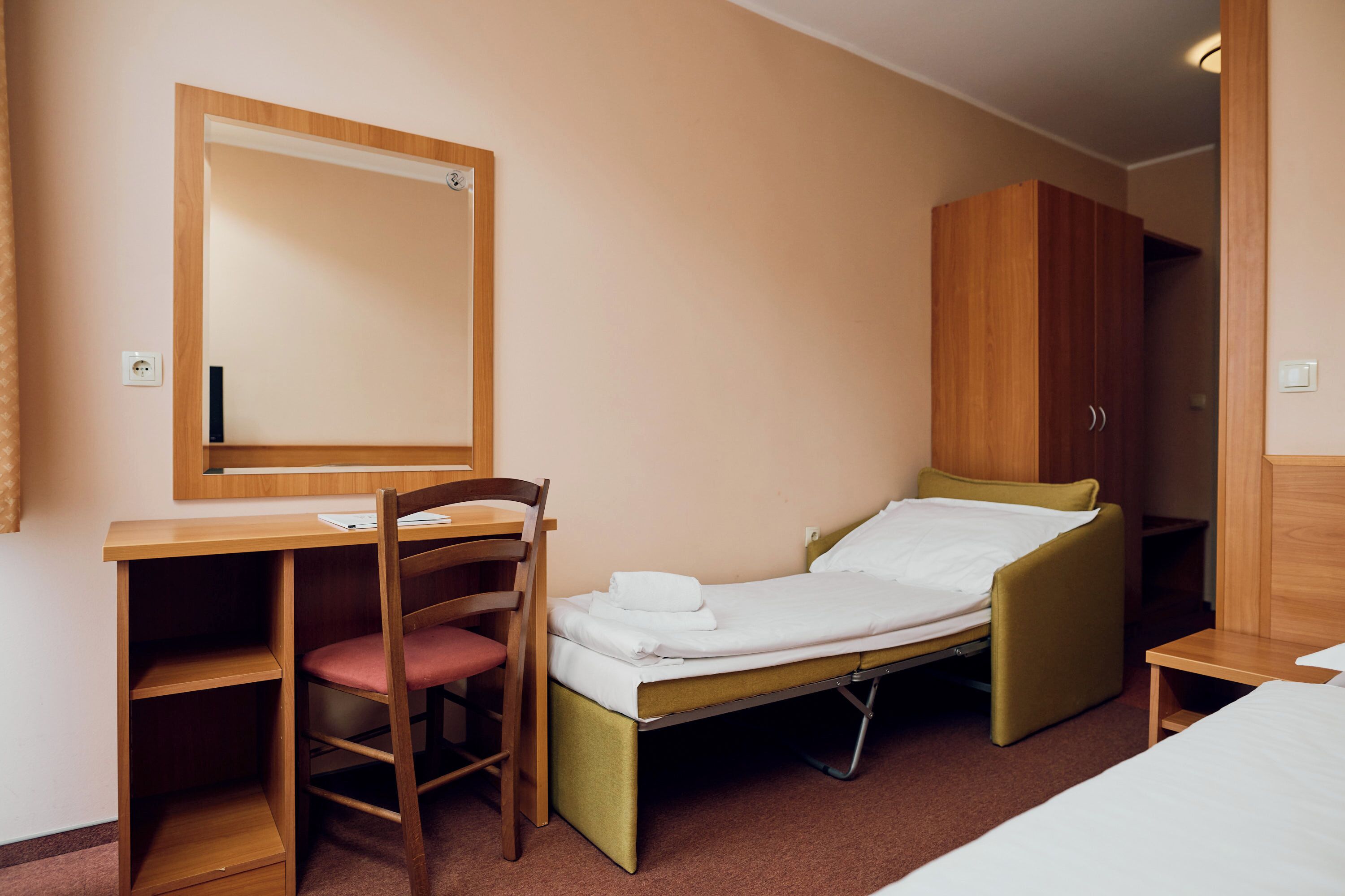 A compact hotel room with a single bed, a desk with a chair, a wardrobe, and a mirror.