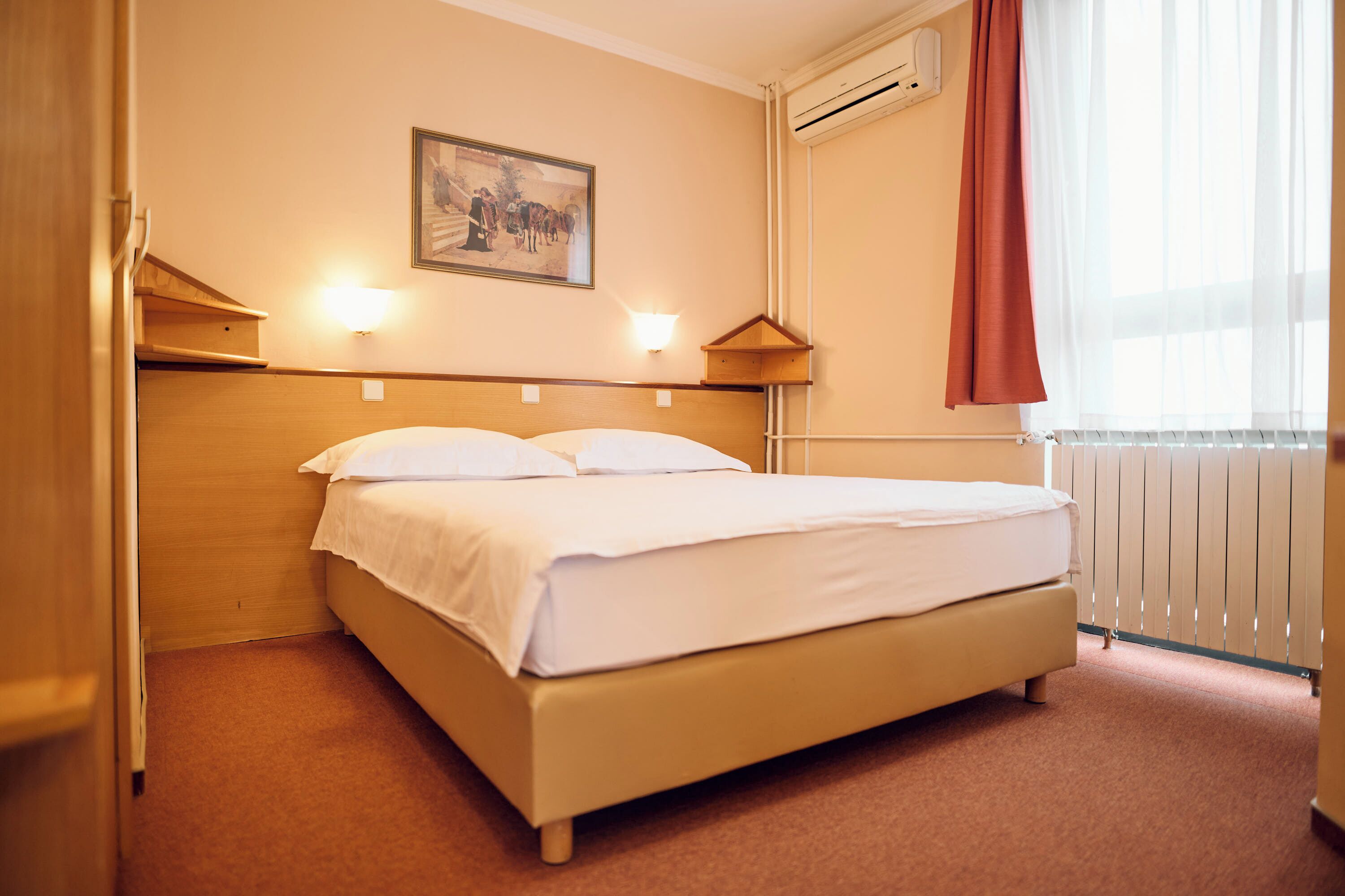 A neatly made bed in a hotel room with a headboard, a wall-mounted picture, and a window with curtains.