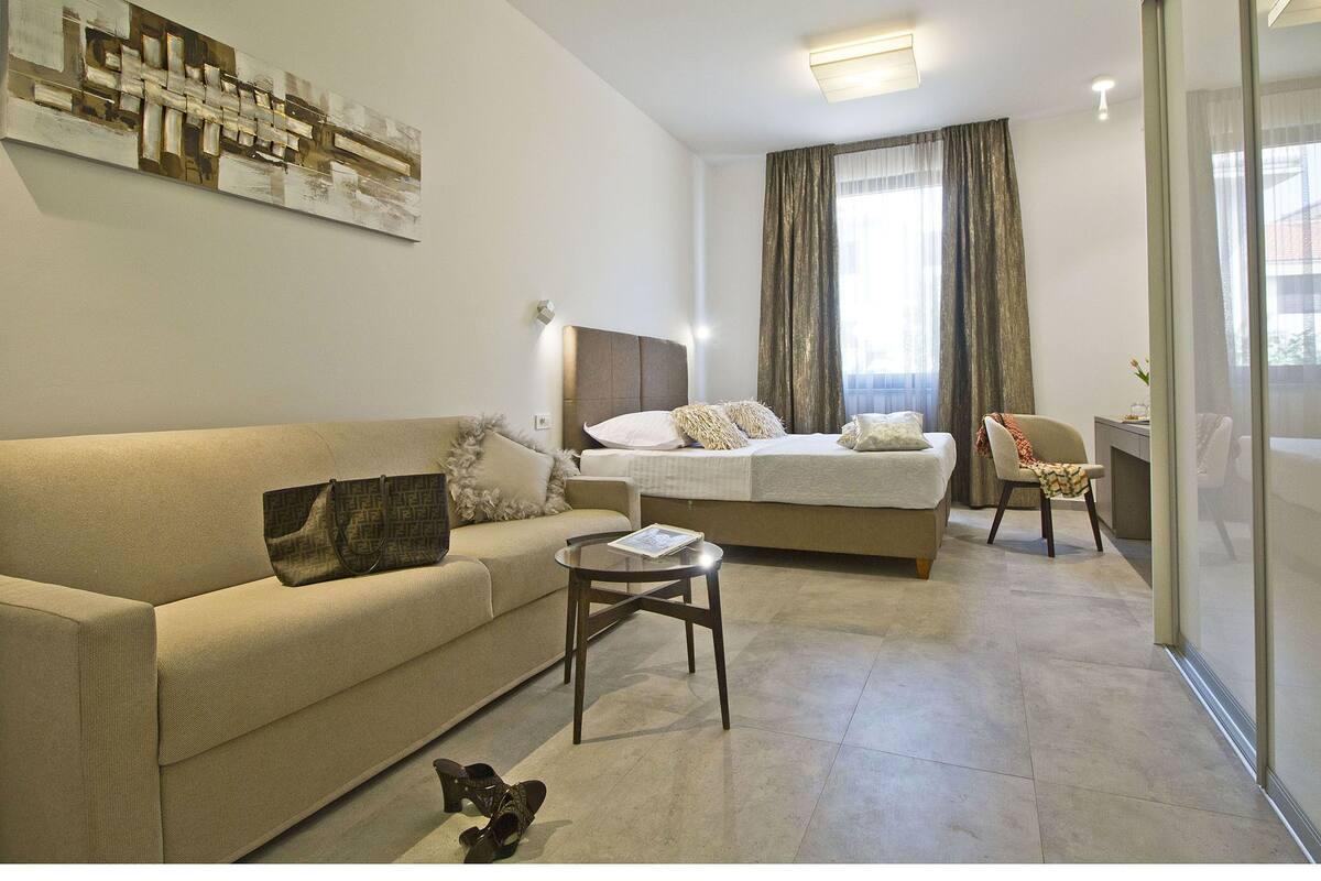 A modern hotel room with a sofa, a bed, a small table, and a glass partition.