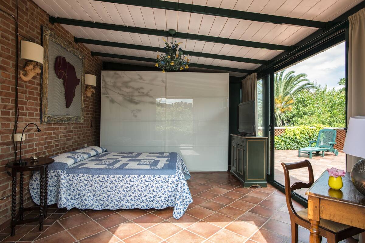 A bedroom with a bed, a TV, a table, and a chair. There is a window with a view of greenery outside.