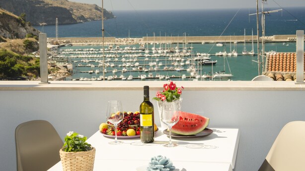 A table with a bottle of wine, a bowl of fruit, a glass of wine, a glass of water, and a slice of watermelon, overlooking a marina with boats. - B&B Nencioli (Castellammare del Golfo)