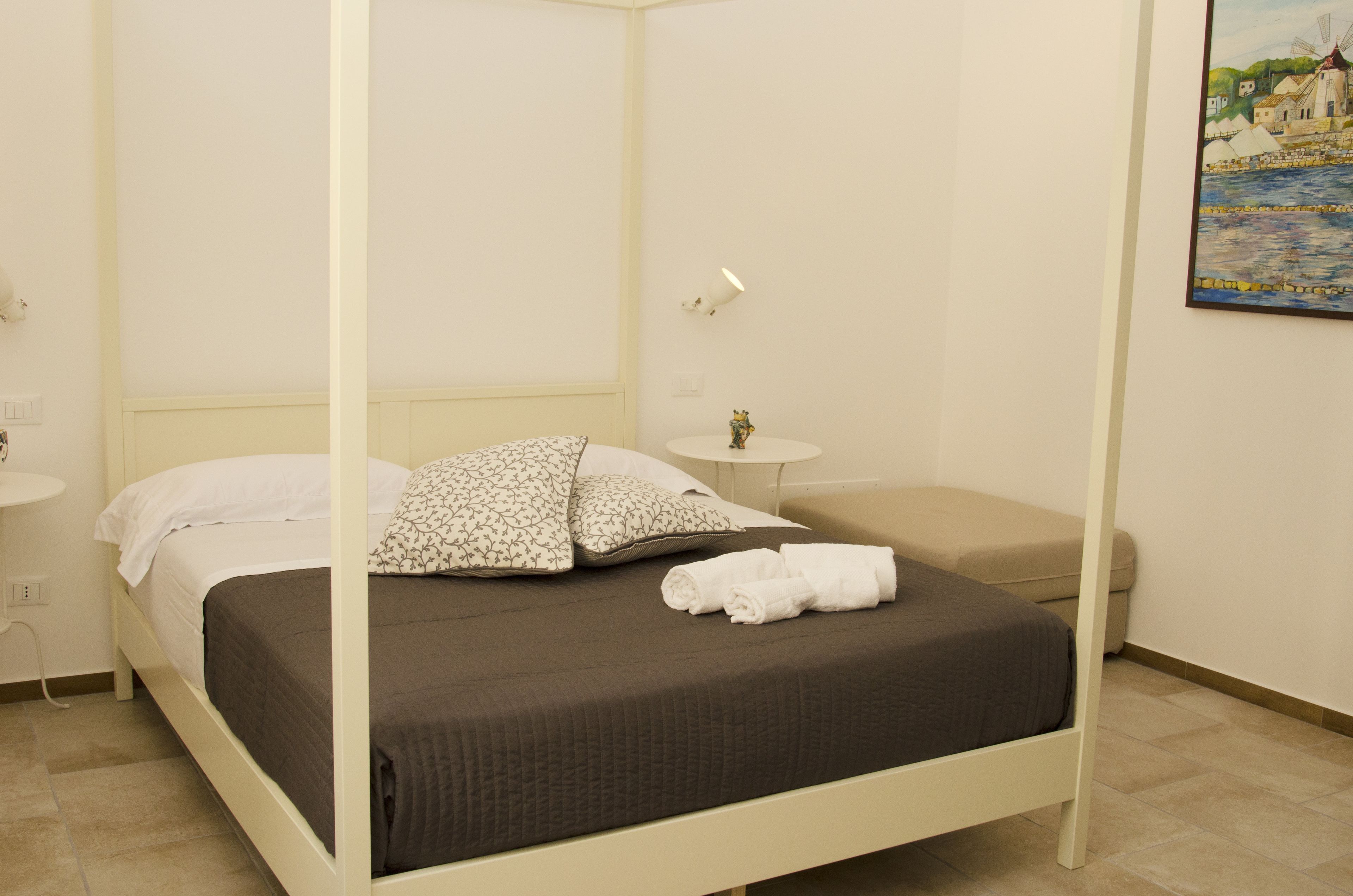 A four-poster bed with a dark brown bedspread and white pillows, a small bedside table with a lamp, and a painting on the wall.