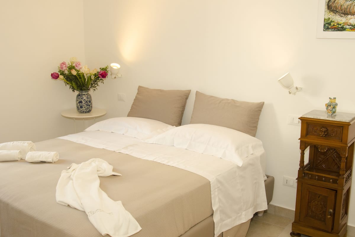 Een net opgemaakt bed met witte lakens, een vaas met bloemen en een houten nachtkastje met een lamp.