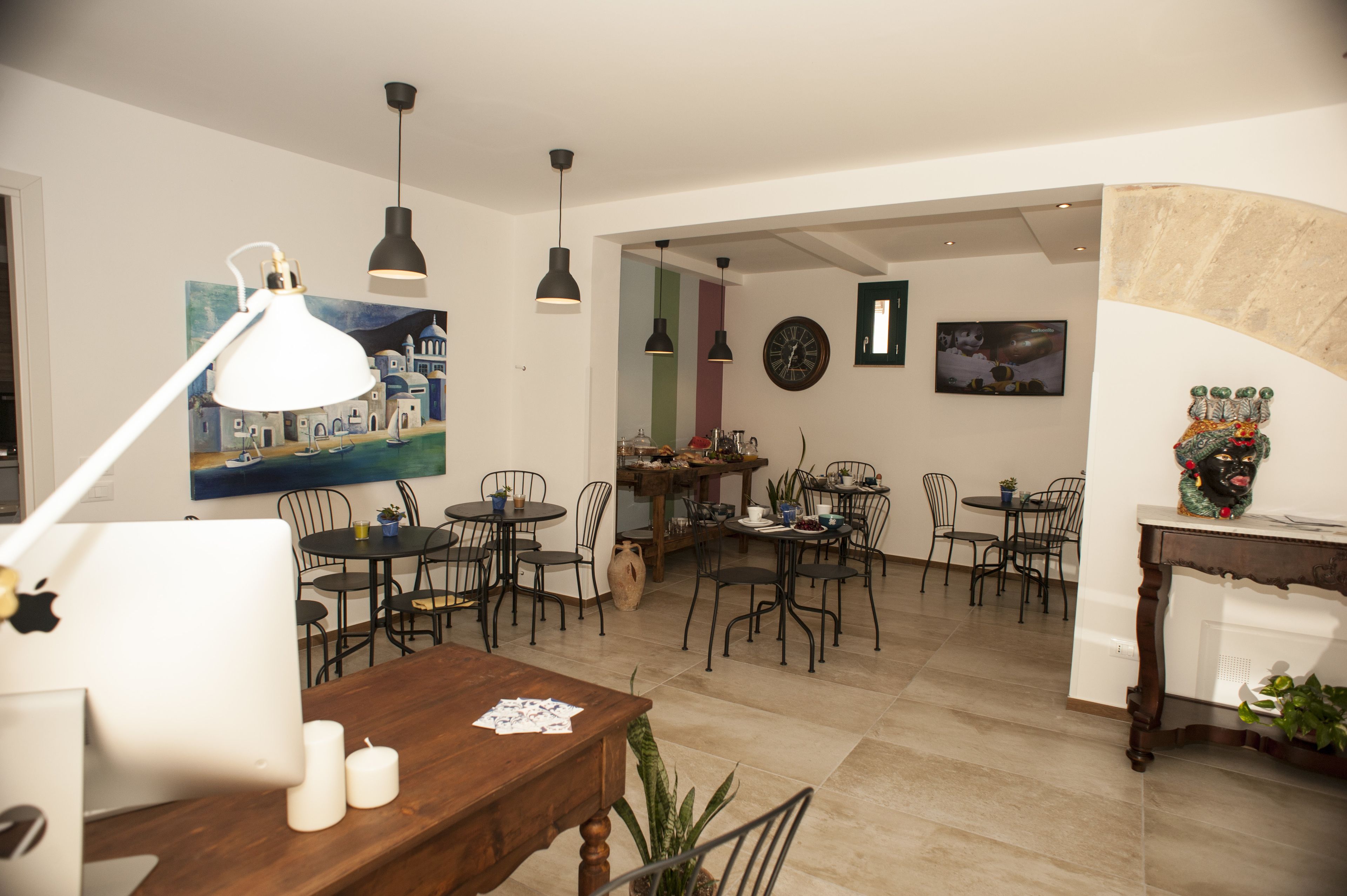 A dining area with multiple tables and chairs, a wooden dining table with a laptop and candles, and various wall decorations including a clock and paintings.