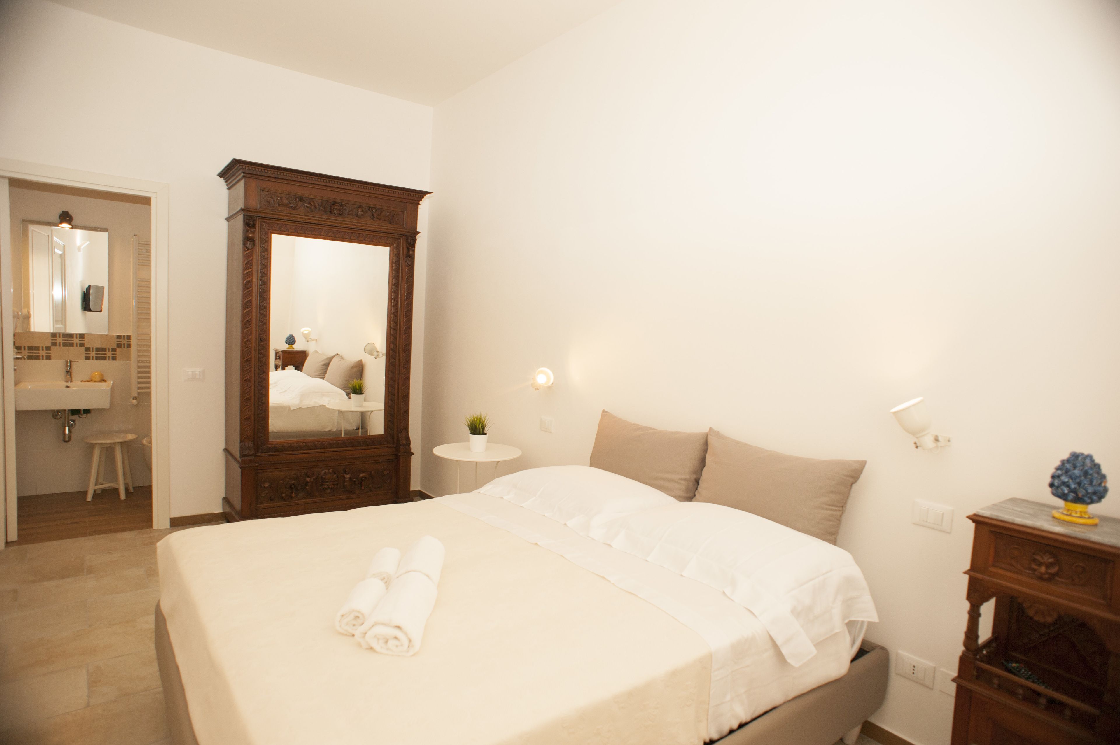 Habitación de hotel con una cama grande, un armario de madera, una mesita redonda con una lámpara y un baño visible a través de una puerta abierta.