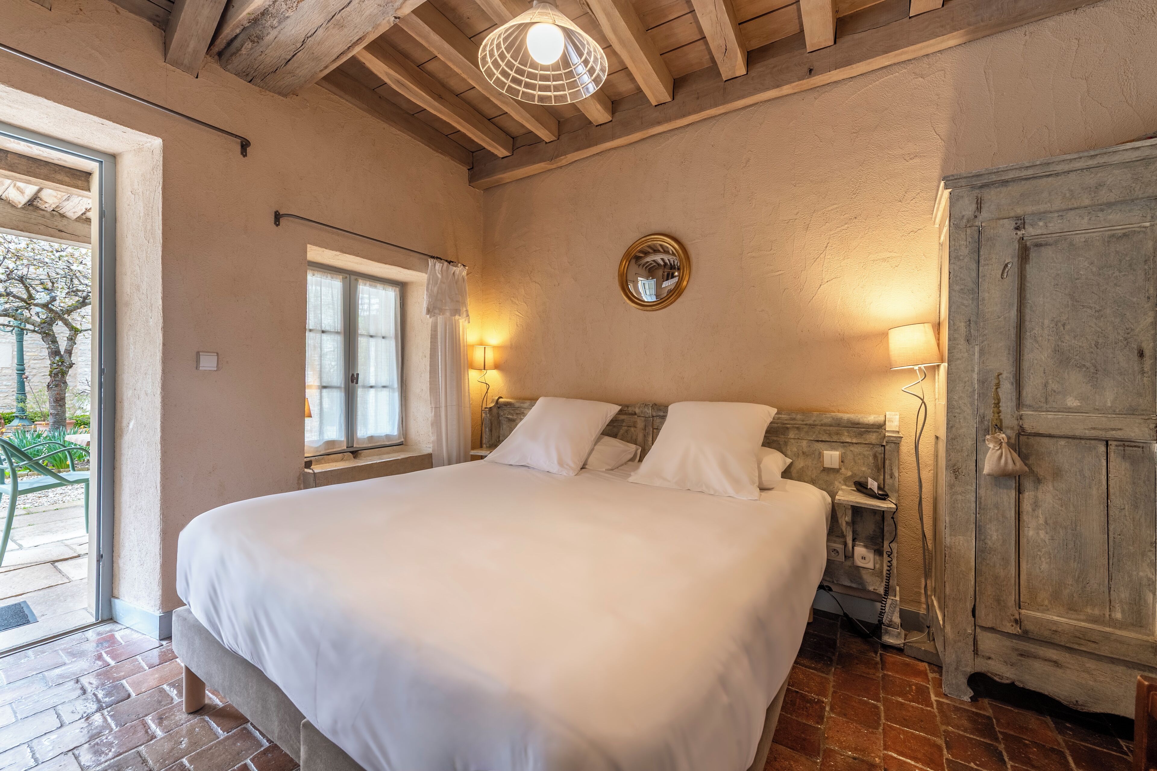 Une chambre avec un grand lit, des poutres apparentes en bois au plafond, une fenêtre avec des rideaux et une porte dotée d’une poignée.