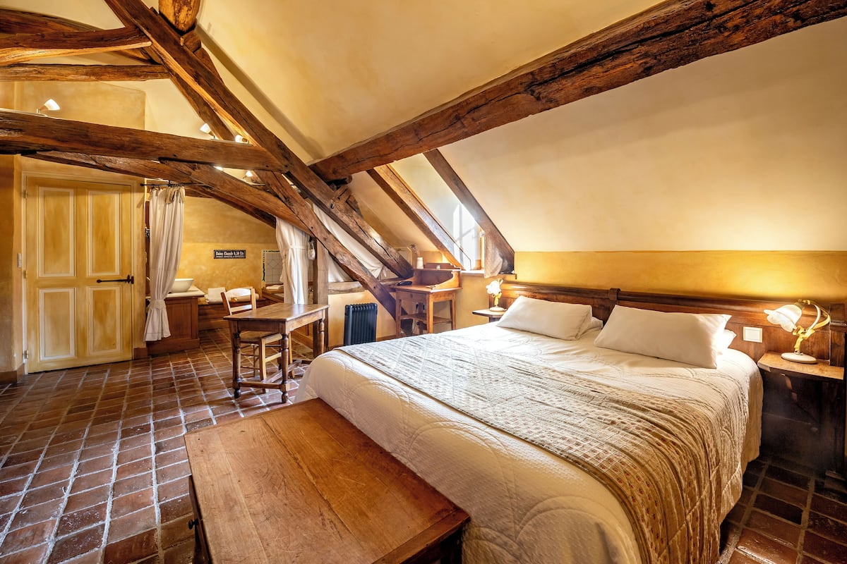 Un dormitorio rústico con vigas de madera a la vista, una cama grande, un banco de madera y una mesita con lámpara.