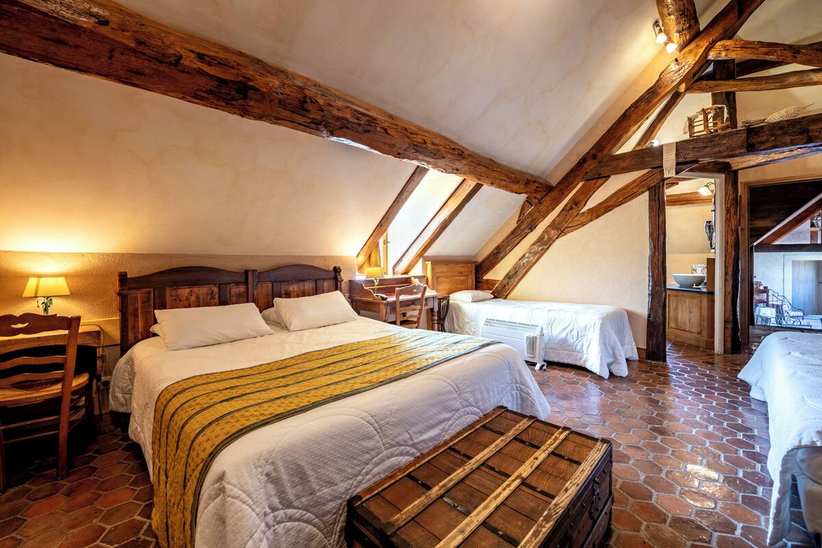Un dormitorio rústico con una cama grande, una cama más pequeña y un cofre de madera. La habitación tiene vigas de madera a la vista y suelo de piedra.