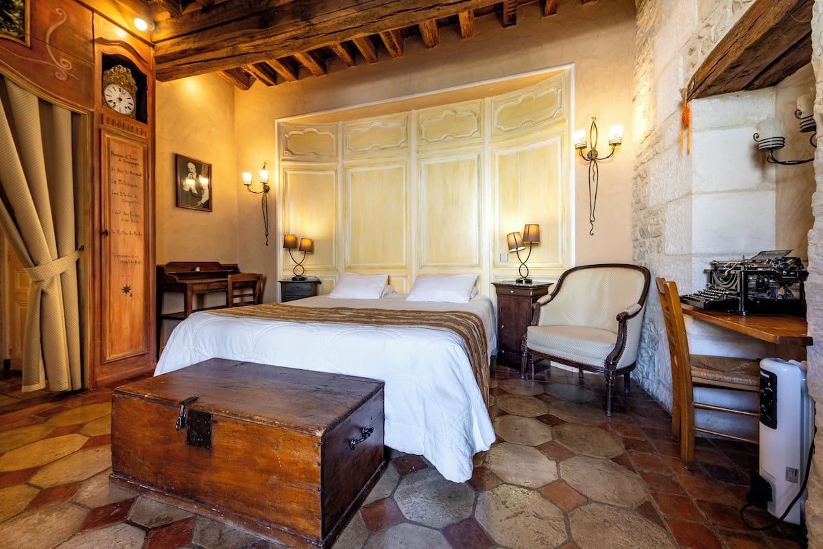 Una habitación tradicional con una cama grande, un cofre de madera, un escritorio con una máquina de escribir y un suelo de baldosas de piedra.