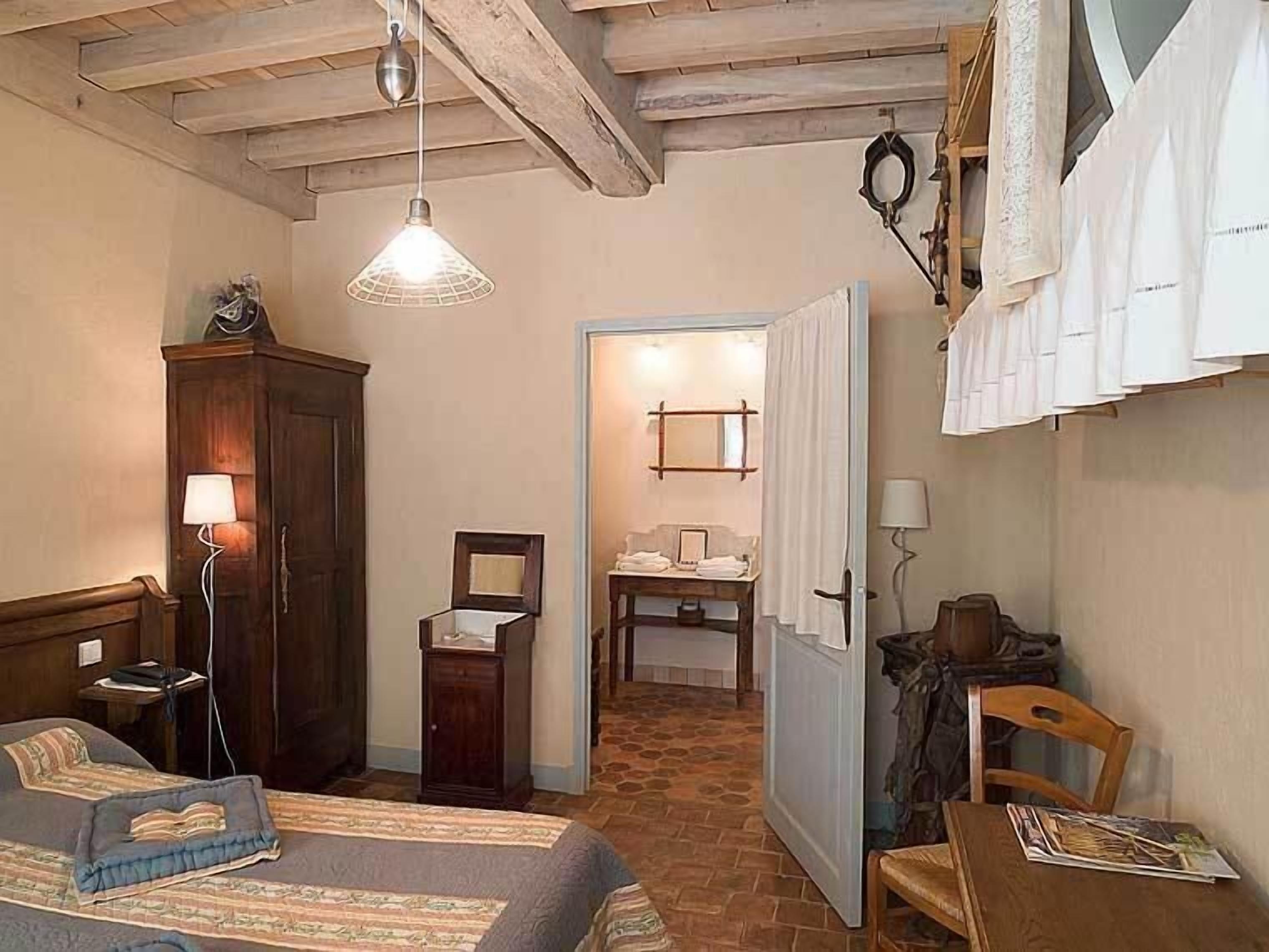 Un dormitorio con cama, un armario de madera, escritorio con silla, lámpara colgante y baño visible a través de una puerta abierta.