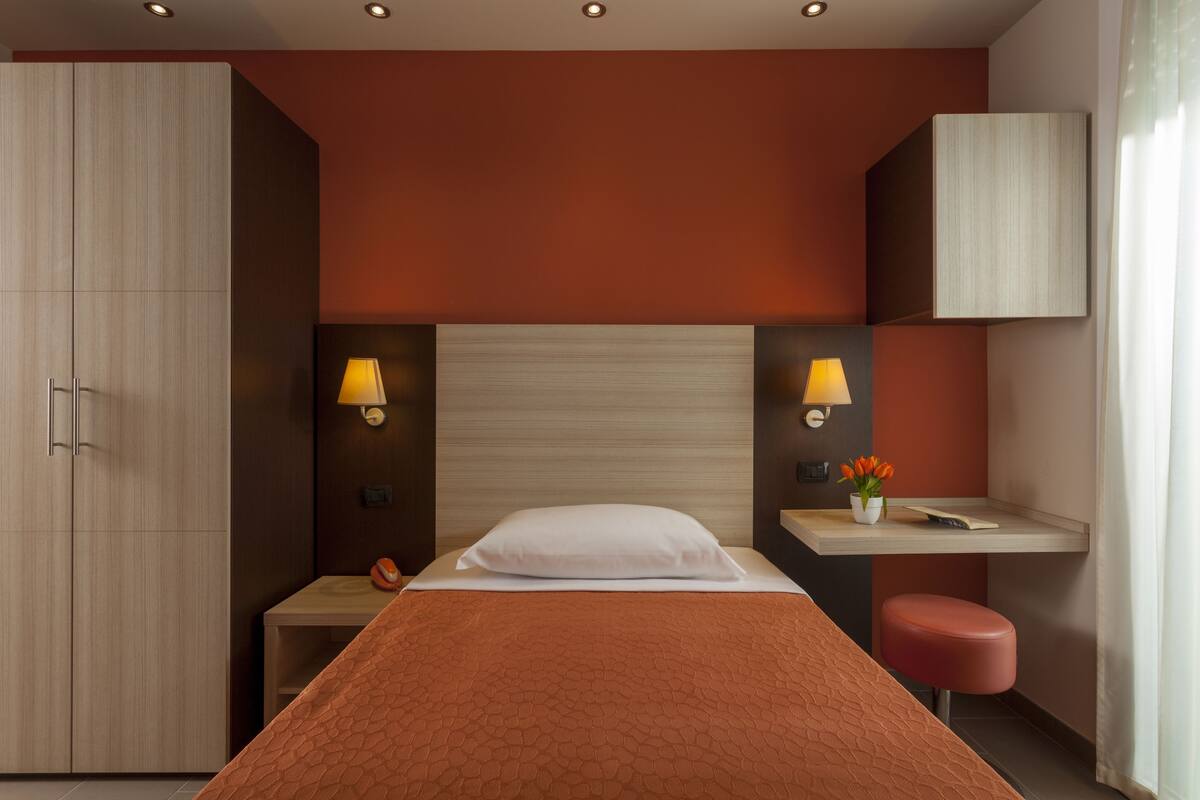 A modern hotel room with a large bed, a desk, a chair, and a wardrobe.