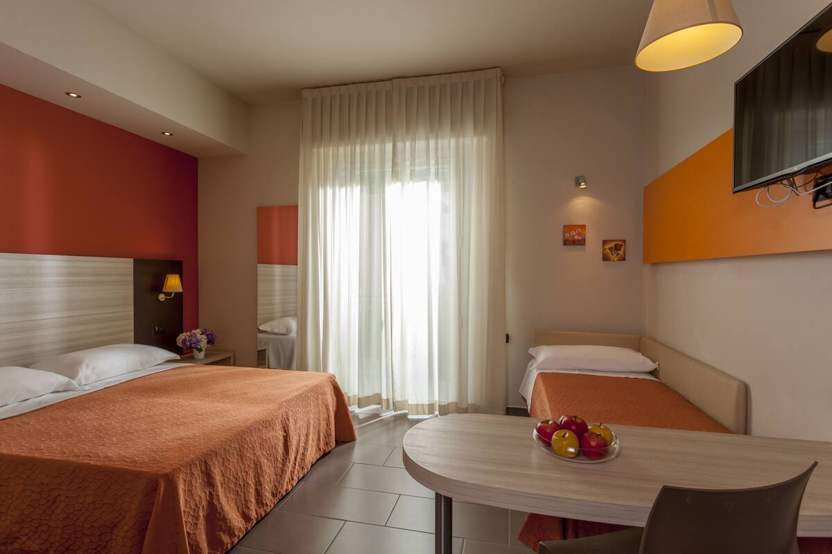 A hotel room with two beds, a dining table with fruit, and a TV mounted on the wall.
