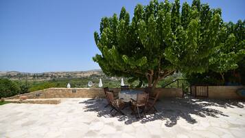 An outdoor patio with stone flooring, a round table, and chairs, surrounded by a stone wall and a tree providing shade.