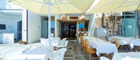 An outdoor dining area with white tables and chairs, covered by yellow umbrellas, and a stone floor.