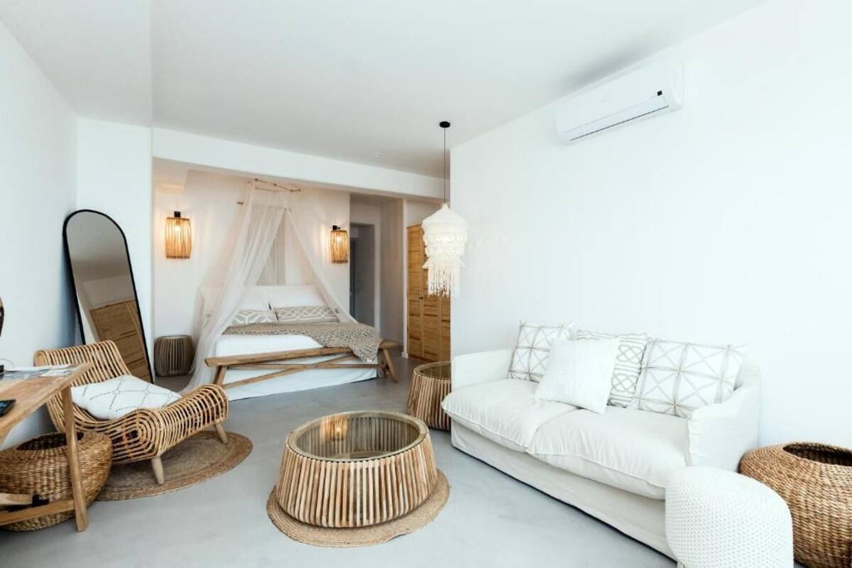 A modern bedroom with a white bed, a sitting area with a white sofa and wicker chairs, a round wicker table, and a large mirror on the wall.