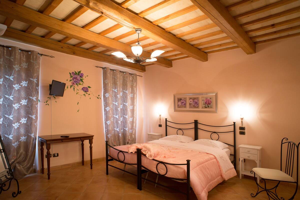 Ein Schlafzimmer mit Holzbalkendecke, einem Bett mit rosa Bettdecke, einem Nachttisch, einem Sessel und einem Schreibtisch.