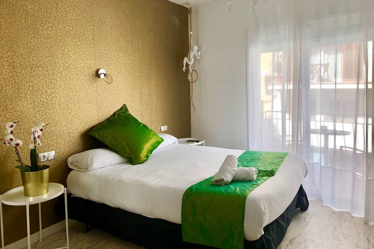 Ein Hotelzimmer mit Bett, einem kleinen Tisch mit Pflanze und Blick durch durchscheinende Vorhänge nach draußen.