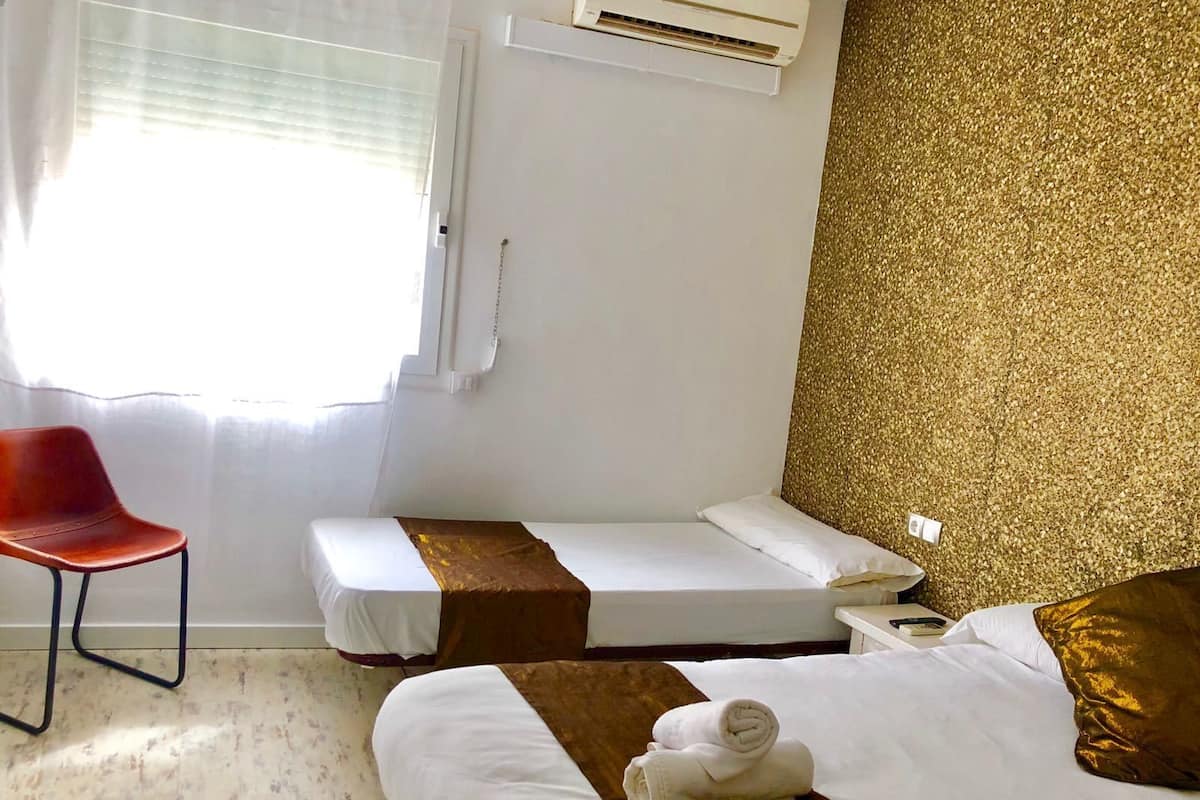 Ein Hotelzimmer mit zwei Einzelbetten, einem roten Sessel, einer Wand mit goldener Struktur und einer Klimaanlage.