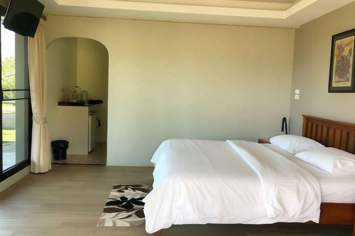 Quarto com cama, cabeceira de madeira, janela com cortinas, mesinha com abajur e quadro pendurado na parede.