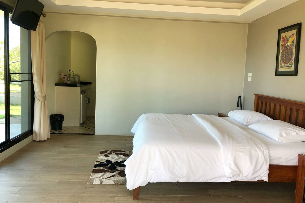 Een slaapkamer met een bed, een houten hoofdbord, een klein nachtkastje, een wandtelevisie en een raam met gordijnen.