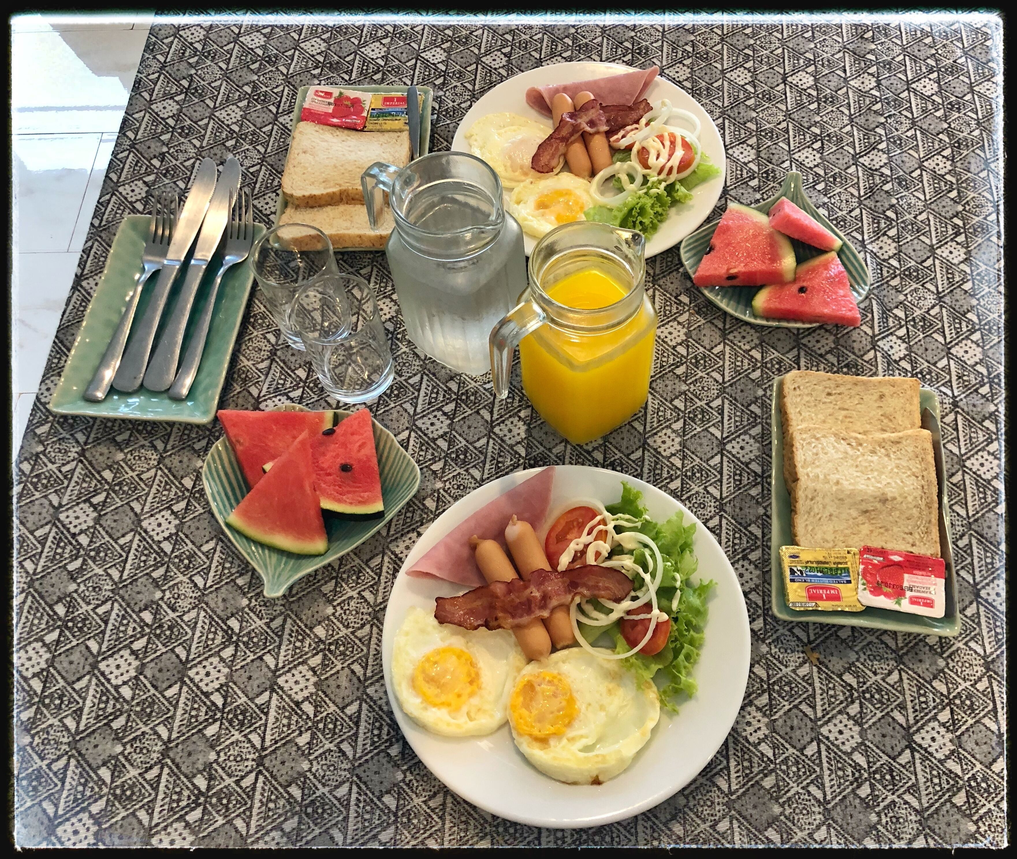 A breakfast table with plates of eggs, bacon, toast, and watermelon, along with a pitcher of orange juice and a glass of water.