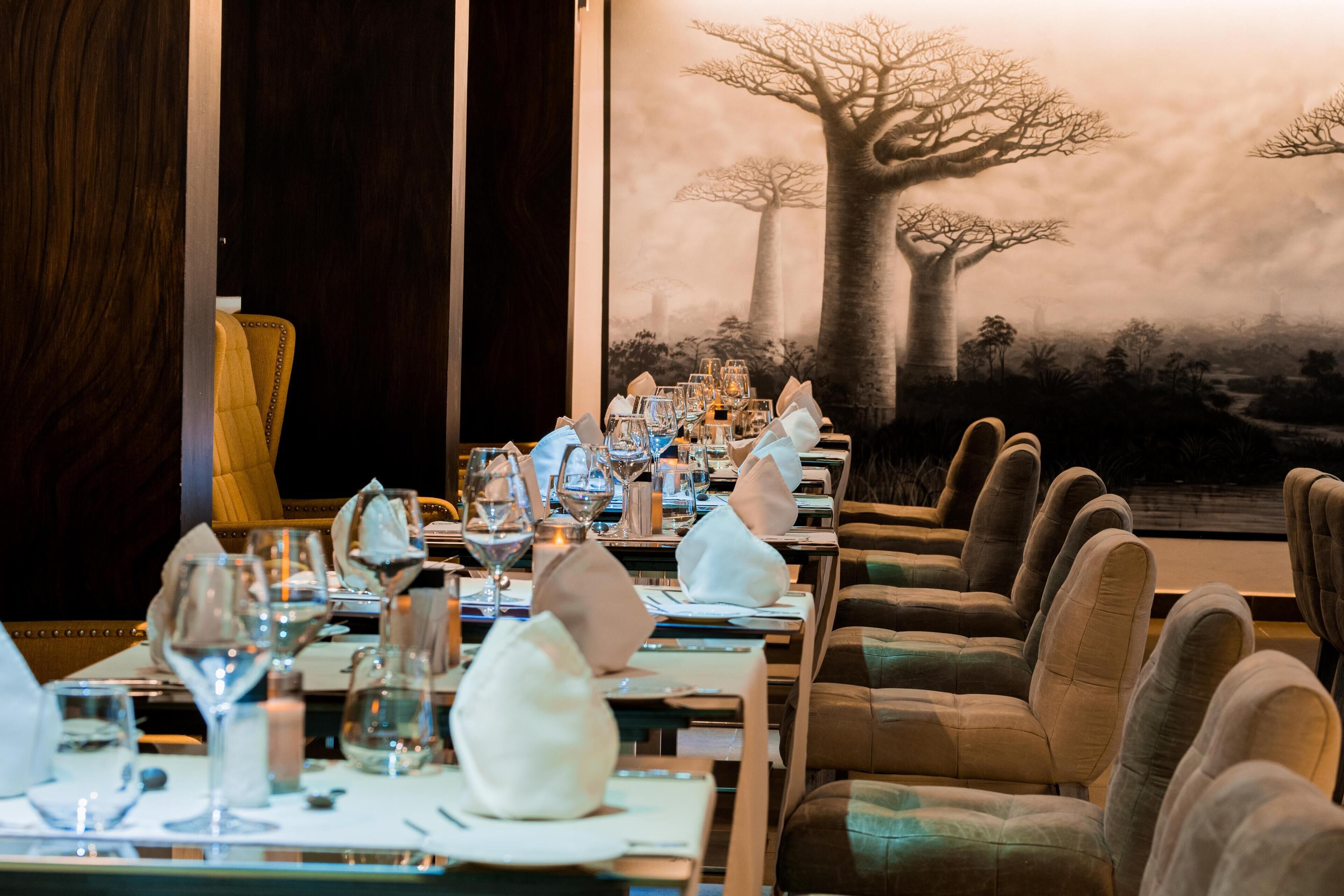 A restaurant with long tables set for dining, a large tree mural on the wall, and leather chairs.