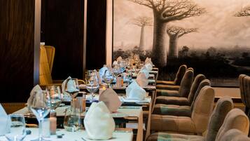 A restaurant with long tables set for dining, a large tree mural on the wall, and leather chairs.