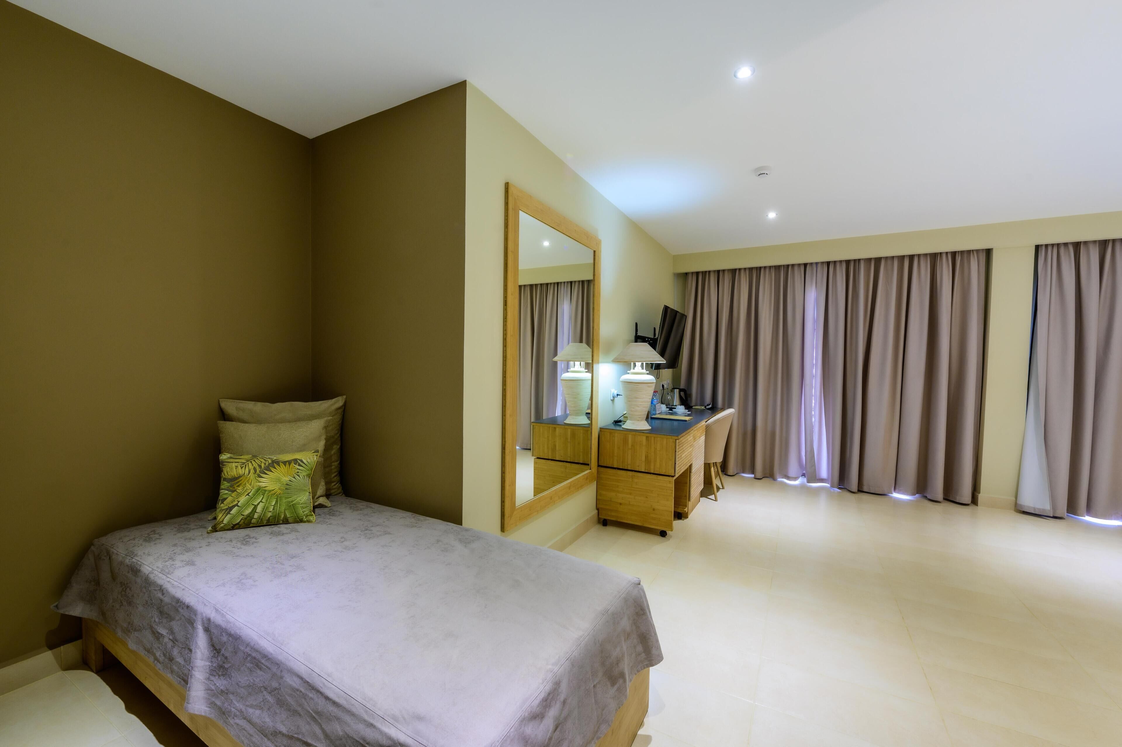 A hotel room with a bed, a desk with a mirror, a chair, and a large window with curtains.