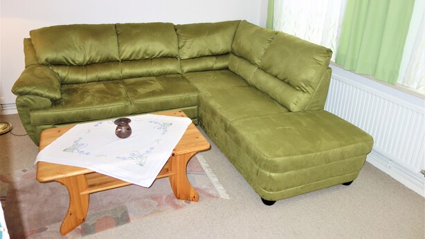 A green sectional sofa with a wooden coffee table in a room with a radiator and a window with green curtains. - Ferienwohnung Tuchtfeld (Goslar)
