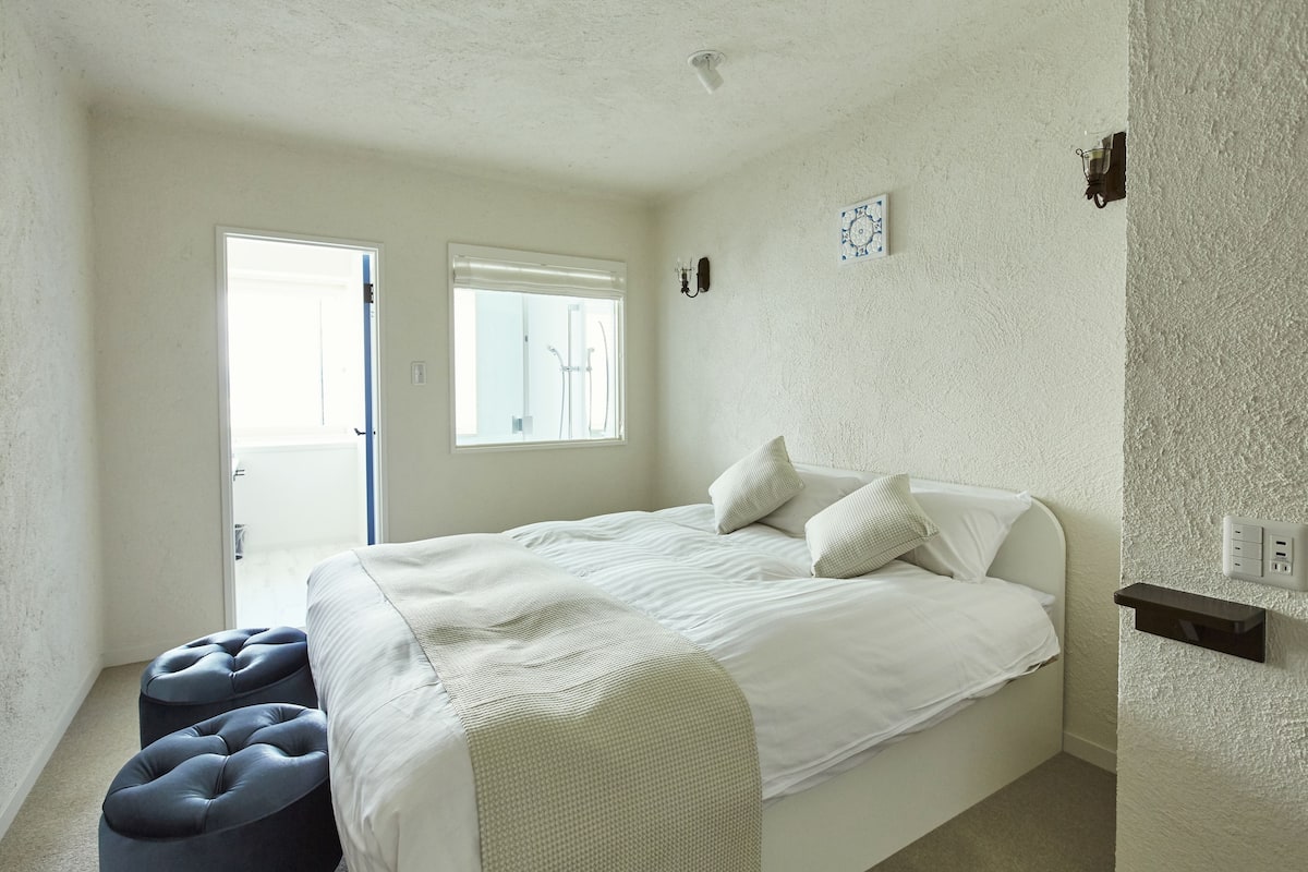 A bedroom with a bed, two ottomans, a window, and a door.