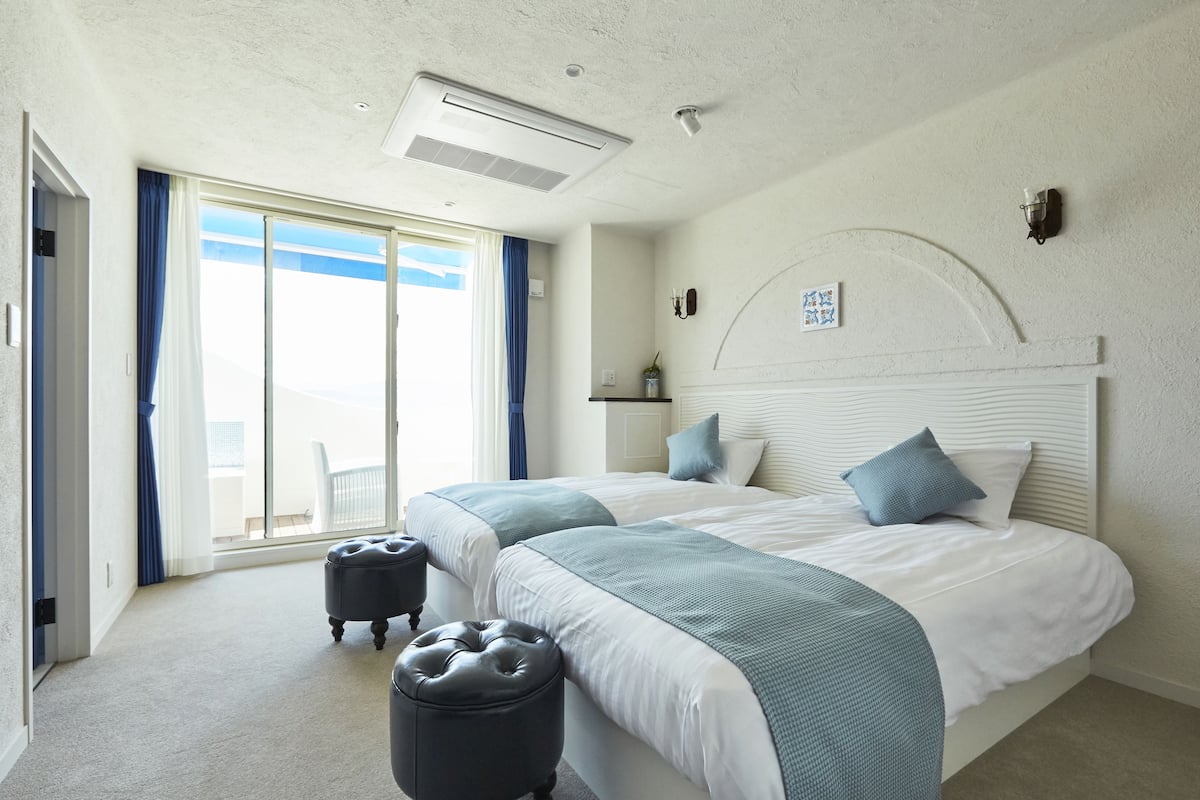 A hotel room with two beds, a large window with curtains, a ceiling fan, and a view of the sky.