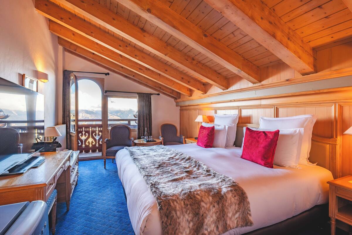Una habitación de hotel con una cama grande, vigas de madera en el techo, un escritorio con computadora y vistas a las montañas.
