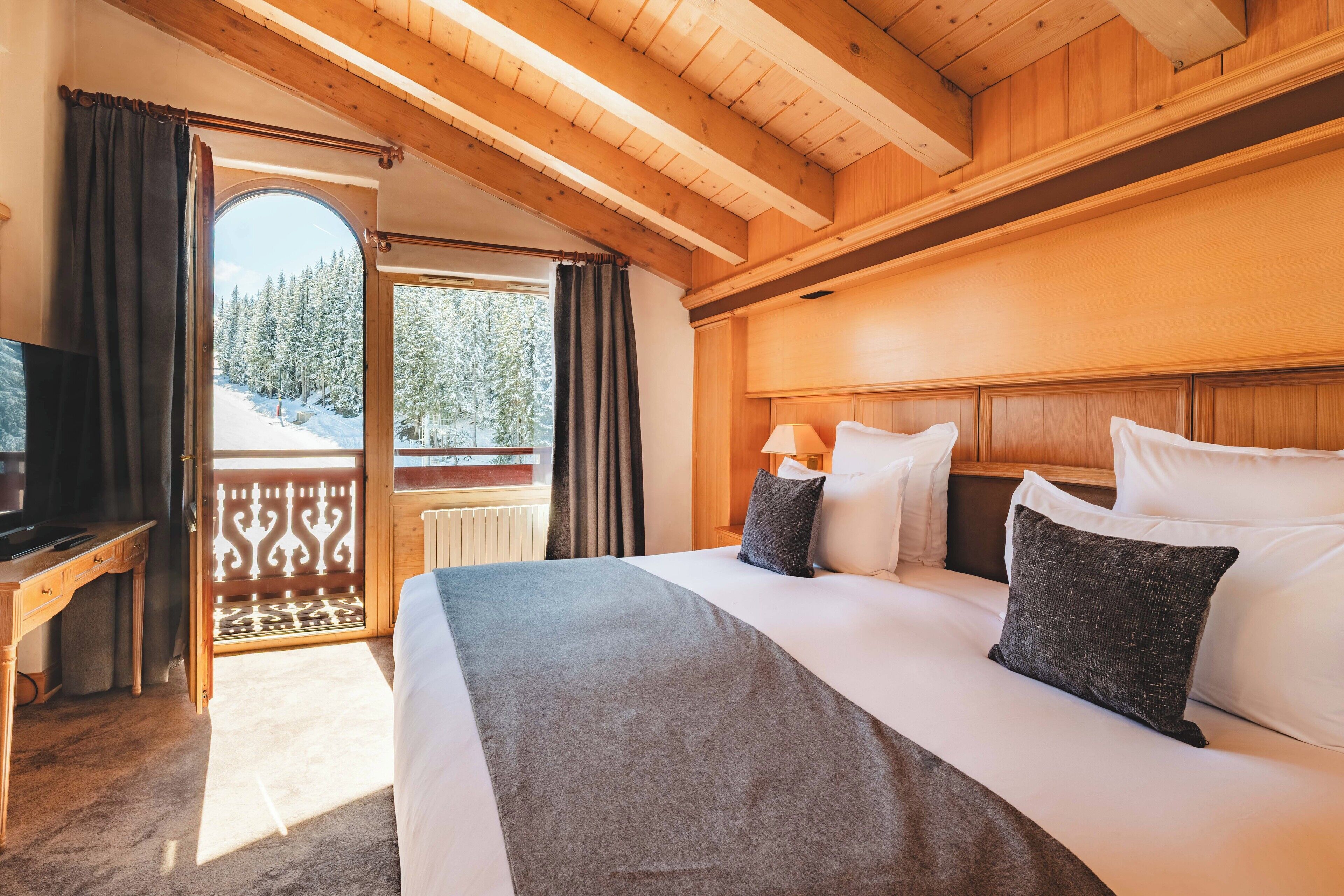 Een houten kamer met een bed, een bureau en uitzicht op een besneeuwd bos door een geopende deur.