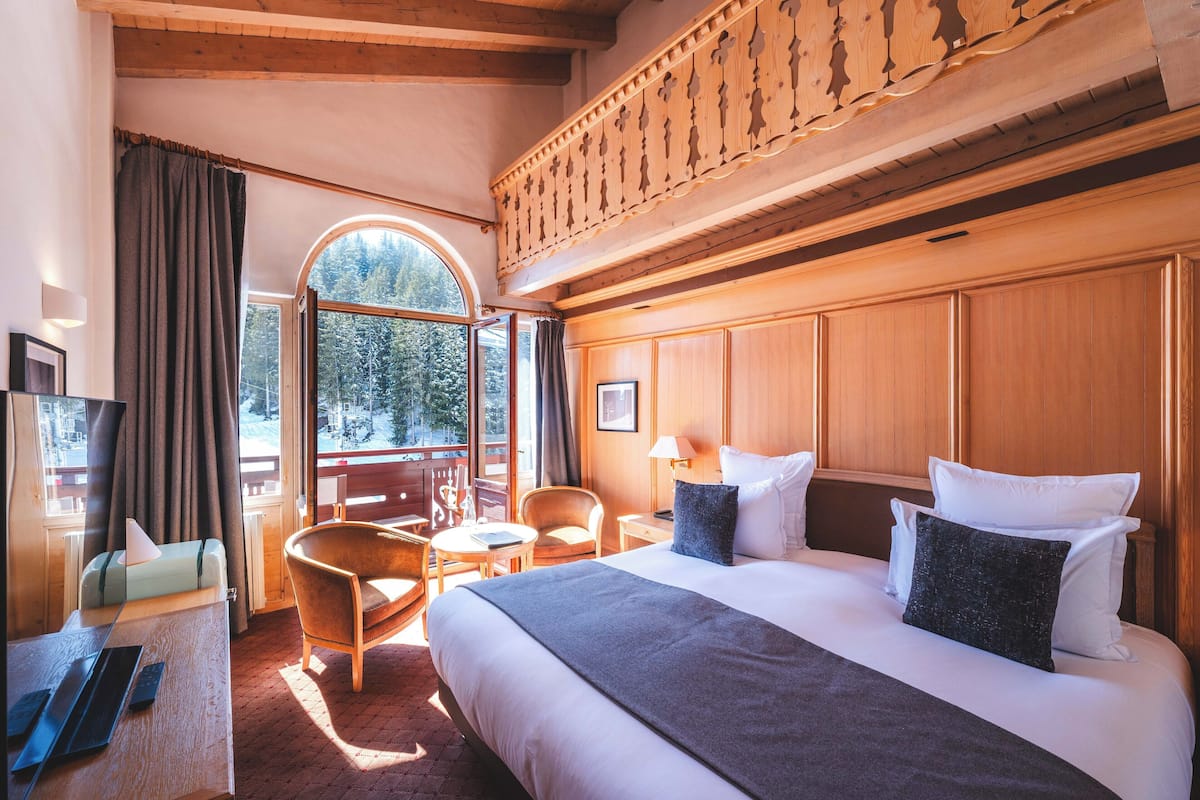 Habitación de hotel con una cama grande, un escritorio con una computadora portátil, una silla y vistas al exterior a través de un ventanal amplio.