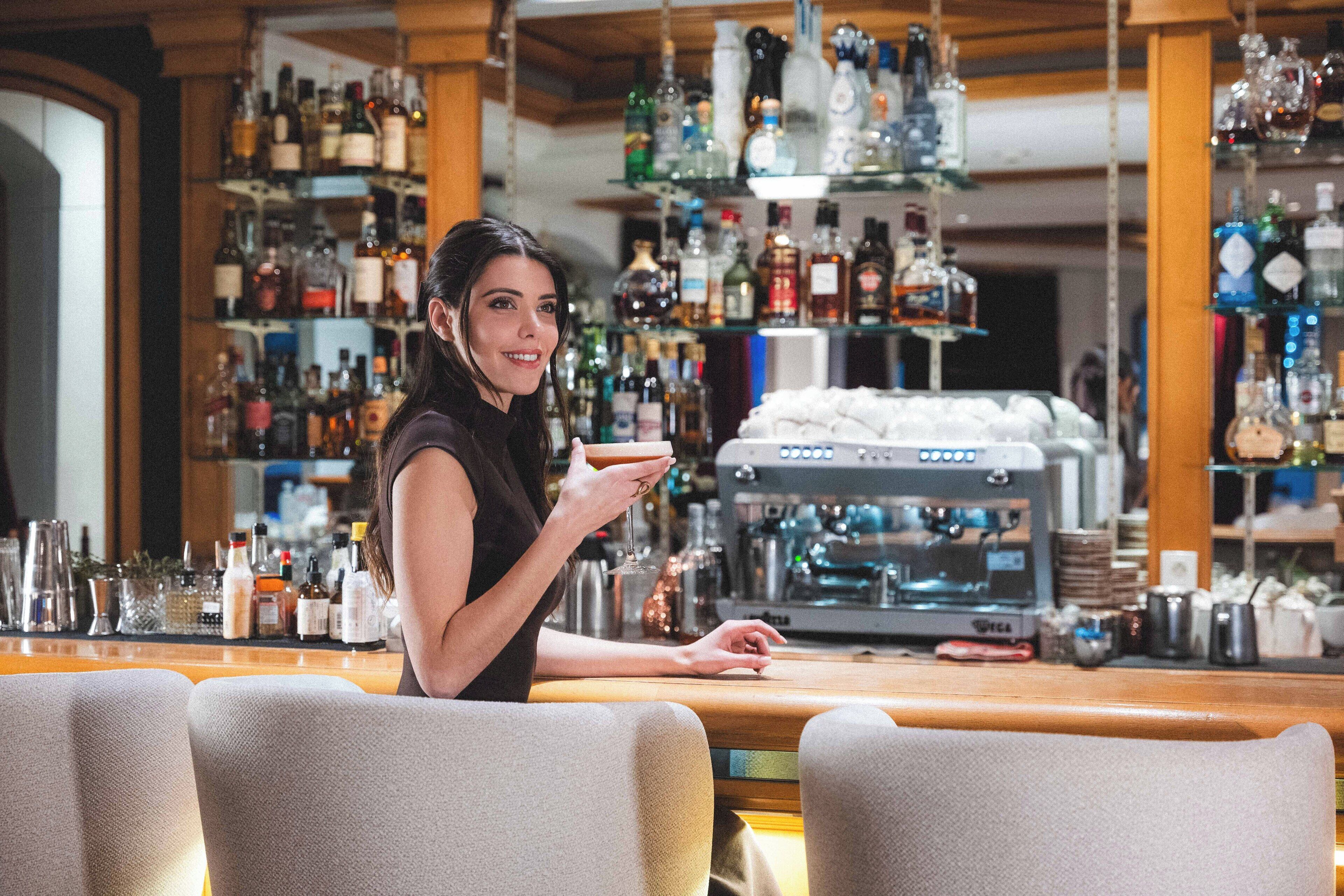 A woman seated at a bar with a variety of liquor bottles in the background.