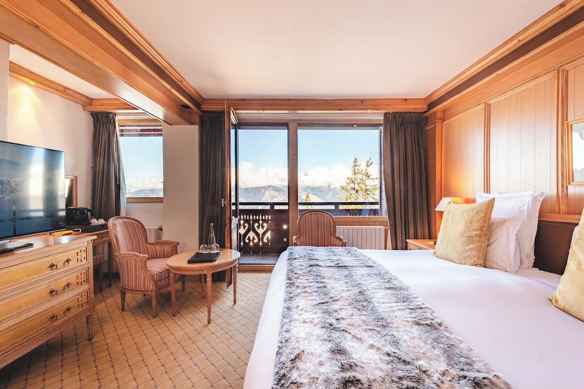 Habitación de hotel con una cama grande, un escritorio con silla, televisión y un balcón con vistas a montañas.
